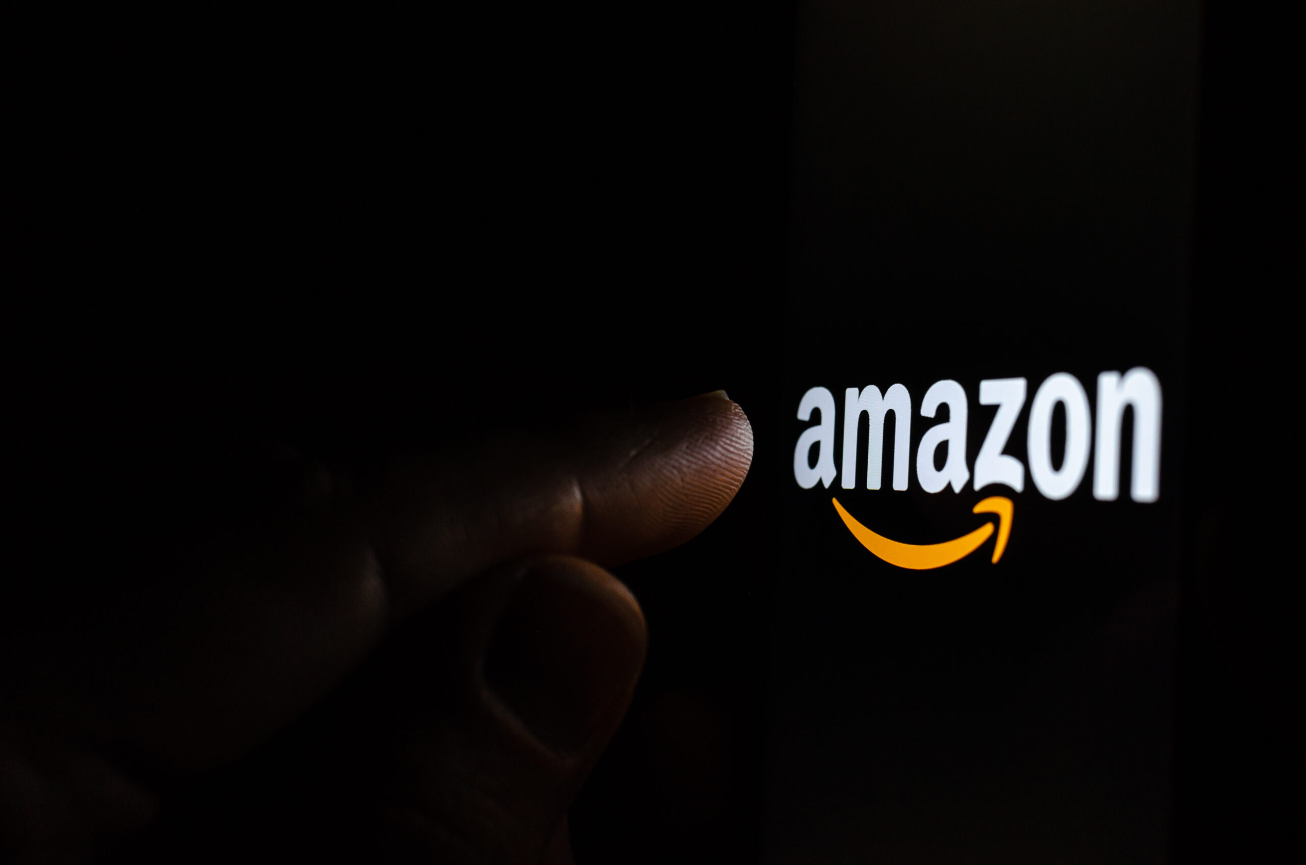 Amazon logo on a smartphone screen in a dark room and a finger pointing at it.