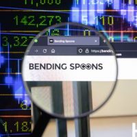 Bending Spoons company logo on a website with blurry stock market developments in the background, seen on a computer screen through a magnifying glass.